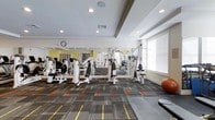 This fitness center offers a comprehensive workout environment designed to accommodate a range of exercise routines. The space is outfitted with multiple white-framed strength training machines arranged across the room, providing users with options for targeting various muscle groups. A large wall mirror spans one side of the space, enhancing the sense of openness while allowing users to monitor their form during workouts. The flooring features a patterned carpet with bold orange, yellow, and grey striping, adding a dynamic visual element to the space. Recessed lighting in the tray ceiling provides consistent illumination throughout, while large windows along one wall are fitted with white horizontal blinds that diffuse ample natural light. A stability ball and additional fitness accessories are visible, indicating the space supports varied workout styles beyond machine-based training. Crown molding along the ceiling adds a refined architectural touch. Built-in shelving is visible along one wall, offering convenient storage for equipment and personal items. The overall layout is open and organized, creating a functional and welcoming fitness environment.