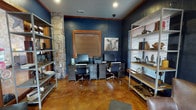 Located in Westworth Village, Texas, this single-floor space blends rustic character with modern functionality through a cohesive and well-appointed design. The interior is defined by deep navy-painted walls accented with warm wood trim, stained concrete flooring in rich amber tones, and a striking natural stone column that anchors the space with texture and architectural interest.

The primary work area features dual dark-toned desks equipped with computers, a printer, and ample drawer storage, flanked on either side by open industrial-style metal shelving. Decorative touches, including Western-themed artwork and curated accessories, reinforce a distinctly Texas aesthetic throughout.

A separate lounge area provides a comfortable retreat with leather armchairs, a small side table, and direct access to an outdoor patio through wood-framed glass French doors fitted with wooden blinds that allow for controlled natural light. A second set of interior French doors with glass panes connects the business center to adjacent spaces, maintaining an open and connected flow.

The overall ambiance is warm yet professional, combining the rugged charm of stone and stained concrete with polished furnishings and thoughtful details. The space is well-suited for both focused work and casual interaction, making it a functional and visually compelling community amenity.