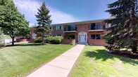 This multi-unit brick building presents a classic and sturdy exterior characterized by warm red brick construction and a well-proportioned facade. A concrete walkway leads from the street directly to the central entrance, which is framed by white trim and a decorative overhead detail, lending a clean and inviting focal point to the front of the building. The upper level features several private balconies with black metal railings, offering residents an outdoor space with views of the surrounding grounds. Large windows are evenly distributed across both levels, allowing for natural light to enter the individual units. The landscaping is carefully maintained, with a broad, neatly trimmed lawn flanking the front walkway, mature evergreen trees providing shade and greenery, and low decorative shrubs lining the building's base. The combination of brick masonry, structured balconies, and verdant outdoor spaces contributes to a tidy and residential atmosphere. The grounds appear spacious and well-kept, enhancing the overall curb appeal of the property. The building's design reflects a functional mid-century aesthetic that balances practicality with a welcoming exterior presence.