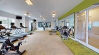 Located at 1421 Baseline Rd in Charlotte, NC, this single-floor fitness center is a well-equipped facility designed to support a comprehensive workout experience. The space is organized into distinct functional zones, each thoughtfully appointed with a variety of equipment to accommodate diverse fitness routines.

The cardio area features a row of treadmills and elliptical machines, all positioned in front of large windows that flood the room with natural light and offer pleasant views of the surrounding greenery. Mounted TVs provide entertainment during workouts. The strength training zone is lined with floor-to-ceiling mirrors along a bold green accent wall, creating a sense of expanded space while allowing users to monitor their form. This area includes a cable machine, adjustable weight benches, and a full dumbbell rack.

A separate studio room offers open floor space with a distinctive green-patterned carpet, spin bikes, and wall art, making it suitable for group classes or individual cycling sessions. Posted fitness center rules and instructional charts ensure a safe and organized environment for all users.

Throughout the facility, a combination of recessed and suspended lighting, ceiling fans, and white walls contribute to a clean, bright, and energizing atmosphere. The layout maximizes usable floor space while maintaining clear pathways between equipment zones.