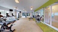 Located at 1421 Baseline Rd in Charlotte, NC, this single-floor fitness center is a well-equipped facility designed to support a comprehensive workout experience. The space is organized into distinct functional zones, each thoughtfully appointed with a variety of equipment to accommodate diverse fitness routines.

The cardio area features a row of treadmills and elliptical machines, all positioned in front of large windows that flood the room with natural light and offer pleasant views of the surrounding greenery. Mounted TVs provide entertainment during workouts. The strength training zone is lined with floor-to-ceiling mirrors along a bold green accent wall, creating a sense of expanded space while allowing users to monitor their form. This area includes a cable machine, adjustable weight benches, and a full dumbbell rack.

A separate studio room offers open floor space with a distinctive green-patterned carpet, spin bikes, and wall art, making it suitable for group classes or individual cycling sessions. Posted fitness center rules and instructional charts ensure a safe and organized environment for all users.

Throughout the facility, a combination of recessed and suspended lighting, ceiling fans, and white walls contribute to a clean, bright, and energizing atmosphere. The layout maximizes usable floor space while maintaining clear pathways between equipment zones.