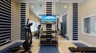Located in Columbia, Tennessee, this fitness center is a thoughtfully designed space built to support a comprehensive workout experience. The room features a striking navy blue and white striped wall treatment with decorative horizontal slat panels that give the space a modern, polished aesthetic. Full-length mirrors are strategically placed throughout, visually expanding the room and allowing users to monitor their form during exercise.

The flooring combines light hardwood with blue rubberized mat sections, offering both durability and comfort across different workout zones. Natural light filters in through large windows fitted with white blinds, creating a bright and energizing atmosphere complemented by recessed ceiling lighting.

The space is outfitted with a well-rounded selection of fitness equipment, including SportsArt treadmills and elliptical trainers for cardiovascular conditioning. A multi-functional cable machine anchors the strength training area, while a full rack of dumbbells spanning a wide weight range provides options for free weight workouts. An adjustable workout bench adds further versatility. A wall-mounted flat-screen television enhances the experience during cardio sessions. The overall ambiance is clean, functional, and motivating — a professional-grade fitness facility designed to meet a variety of fitness needs.