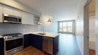 This contemporary single-floor residence is designed with a clean, modern aesthetic and an efficient open-concept layout. The kitchen serves as the centerpiece of the living space, showcasing bold two-tone cabinetry — navy blue on the lower cabinets and crisp white on the upper — complemented by a gray subway tile backsplash and a white quartz peninsula countertop. A stainless steel undermount sink with a pull-down faucet, a gas range, built-in microwave, and a full-size refrigerator round out the well-appointed kitchen. Pendant lights with cone-shaped shades and a track lighting system provide layered illumination throughout the open area. The living space flows naturally from the kitchen, featuring warm hardwood-style flooring that runs throughout the unit. Large floor-to-ceiling windows and a wide sliding glass door flood the living area with natural light and offer views of the surrounding urban landscape. A bedroom with an oversized picture window provides additional natural light and treetop views. The bathroom includes a clean, white soaking tub and shower surround. A stacked washer and dryer unit is conveniently integrated into the layout, adding practical in-unit laundry access. The overall design is cohesive, contemporary, and thoughtfully appointed.