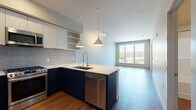 This contemporary single-floor residence is designed with a clean, modern aesthetic and an efficient open-concept layout. The kitchen serves as the centerpiece of the living space, showcasing bold two-tone cabinetry — navy blue on the lower cabinets and crisp white on the upper — complemented by a gray subway tile backsplash and a white quartz peninsula countertop. A stainless steel undermount sink with a pull-down faucet, a gas range, built-in microwave, and a full-size refrigerator round out the well-appointed kitchen. Pendant lights with cone-shaped shades and a track lighting system provide layered illumination throughout the open area. The living space flows naturally from the kitchen, featuring warm hardwood-style flooring that runs throughout the unit. Large floor-to-ceiling windows and a wide sliding glass door flood the living area with natural light and offer views of the surrounding urban landscape. A bedroom with an oversized picture window provides additional natural light and treetop views. The bathroom includes a clean, white soaking tub and shower surround. A stacked washer and dryer unit is conveniently integrated into the layout, adding practical in-unit laundry access. The overall design is cohesive, contemporary, and thoughtfully appointed.