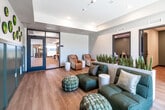Located at 425 SW 11th St in Des Moines, Iowa, this thoughtfully designed wellness suite offers a calm, professional environment across a single floor. The main living area serves as a lounge and reception space, immediately setting a tranquil tone with warm hardwood-style flooring, recessed lighting, and clean white ceilings. A striking accent wall features wainscoting in a muted sage green, adorned with a curated arrangement of circular preserved moss art pieces, adding a biophilic and artistic focal point. Lush snake plants serve as a natural room divider, complementing the organic aesthetic. Comfortable modular seating in deep teal and woven ottoman poufs create an inviting gathering area, while a window offers views of the surrounding Des Moines cityscape. A set of massage chairs and a glass-paneled door leading to an adjacent dining or community area add functional versatility to the lounge. A private consultation room features neutral gray tones, light wood flooring, a plush barrel chair, and framed abstract wall art, providing a quiet and intimate setting. The treatment room is equipped with a built-in countertop, a utility sink, light wood cabinetry with modern black hardware, a white subway tile backsplash, and a sage green accent wall, offering a clean and clinical yet welcoming environment for care.