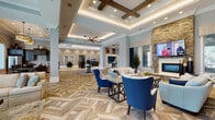 This elegantly designed single-floor community clubhouse offers a seamlessly connected series of social and functional spaces, each thoughtfully appointed with high-quality finishes and comfortable furnishings. The centerpiece of the facility is a grand open lounge area distinguished by dramatic coffered ceilings with integrated ambient lighting, decorative wood beam accents, and a striking herringbone-patterned hardwood floor. Plush seating arrangements in neutral and navy tones invite relaxed conversation, while a stacked-stone accent wall housing a fireplace and mounted television adds warmth and visual interest to the space. Large windows dressed with tailored valances and drapes throughout the facility allow generous natural light and views of the surrounding community. A dedicated sitting area features paired armchairs and full sofas arranged around a wood coffee table, offering a quiet retreat. The kitchen and dining area showcases rich dark cabinetry, a mosaic tile backsplash, stainless steel appliances, and earthy natural stone tile flooring, complemented by a casual café-style dining nook with modern bistro tables and banquette seating. A business center nook provides workstation computers set against a marble-effect feature wall, combining practicality with refined aesthetics. Every area reflects a consistent, upscale design sensibility throughout.
