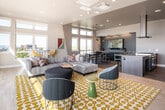 This thoughtfully designed single-floor amenity space combines contemporary style with functional comfort across several distinct yet interconnected areas. The open-plan layout is anchored by a spacious lounge featuring a large sectional sofa, accent chairs, and a bold patterned area rug in gold and white, all set atop warm hardwood flooring. Abundant natural light pours in through oversized windows and full-height glass sliding doors that connect seamlessly to an outdoor terrace, offering open views of the surrounding neighborhood and skyline.

The gourmet kitchen is a standout feature, centered around a generous island with a smooth quartz countertop and seating for multiple guests via upholstered green bar stools. A professional-grade gas range with a sleek overhead hood, dark tile backsplash, rich wood cabinetry, and a stainless steel refrigerator complete the well-appointed cooking space.

A dedicated conference or meeting area provides a more focused setting, furnished with a wood dining table, mixed seating, a wall-mounted display screen, and teal-toned built-in cabinetry. Clerestory windows flood this room with natural light while maintaining a sense of privacy. Throughout the space, recessed lighting, contemporary artwork, and a cohesive neutral-and-teal color palette contribute to a polished, inviting atmosphere suited for both social gatherings and productive meetings.