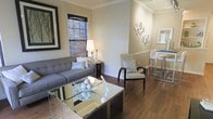 This well-appointed single-floor space offers a thoughtfully designed interior that balances comfort and style. The living area serves as the heart of the home, featuring warm hardwood floors that extend throughout and neutral-toned walls accented by classic crown molding. A plush gray sofa anchors the seating area, complemented by a sleek mirrored coffee table that adds a touch of contemporary elegance. A cushioned armchair with dark wood frame provides additional seating and a refined accent to the room. Natural light filters in through a bamboo-shaded window, lending a soft, inviting ambiance to the space. Decorative wall art and a set of circular mirrored wall accents add personality and visual interest to the room. The living area flows naturally into a dining space, where a high-top table with matching chairs creates a casual yet sophisticated setting. A framed bulletin board or artwork on the adjacent wall adds a functional decorative touch. The overall design palette — warm neutrals, rich wood tones, and metallic accents — creates a cohesive and welcoming atmosphere throughout this single-level home.