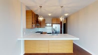 This single-floor residence showcases a contemporary kitchen that serves as the heart of the space. The kitchen is defined by rich, warm-toned wood cabinetry that lines the walls and extends to the ceiling, offering generous storage while contributing a natural, earthy aesthetic. A spacious kitchen island with a clean white quartz countertop and light wood paneling on its base provides ample workspace and a natural gathering point. Twin globe-style pendant lights with brass accents hang above the island, casting a warm, sophisticated glow throughout the space. The backsplash features a subtle mosaic tile design that adds texture and visual interest without overwhelming the overall palette. Stainless steel appliances, including a gas range and a large French door refrigerator, lend a polished, professional-grade feel to the culinary space. Recessed lighting complements the pendant fixtures, ensuring the kitchen is well-lit throughout. The flooring transitions from rich dark hardwood to the kitchen area seamlessly, tying the overall design together. The clean lines, neutral tones, and high-quality finishes throughout create a cohesive and functional environment that balances modern design sensibilities with everyday practicality.