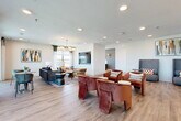 This thoughtfully designed community space in Allen, Texas offers a single-floor open-plan layout that seamlessly connects multiple social zones. The interior is anchored by warm wood-look flooring that runs throughout, complemented by crisp white walls and recessed lighting that maintain a bright, welcoming atmosphere.

The lounge area is generously proportioned, featuring multiple seating arrangements including plush rust-colored velvet armchairs, high-back gray upholstered sofas, and a cozy sitting nook outfitted with layered rugs and a multi-drum chandelier. Large windows along the perimeter flood the space with natural light and frame sweeping outdoor views of the Allen, TX landscape, enhancing the open and airy feel.

A standout kitchenette anchors one end of the space, finished with soft teal cabinetry, white quartz countertops, a sleek matte-black faucet, and a dramatic stainless steel range hood. An eye-catching array of brass pendant lights crowns a large central island with leather barstool seating, making it an ideal spot for casual gatherings.

Curated artwork, including bold abstract canvases in warm earth tones and jewel-toned blues, adds personality and visual interest throughout. A wall-mounted TV and defined seating clusters create purposeful zones within the open layout. The overall ambiance is modern, sophisticated, and inviting.