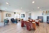 This thoughtfully designed community space in Allen, Texas offers a single-floor open-plan layout that seamlessly connects multiple social zones. The interior is anchored by warm wood-look flooring that runs throughout, complemented by crisp white walls and recessed lighting that maintain a bright, welcoming atmosphere.

The lounge area is generously proportioned, featuring multiple seating arrangements including plush rust-colored velvet armchairs, high-back gray upholstered sofas, and a cozy sitting nook outfitted with layered rugs and a multi-drum chandelier. Large windows along the perimeter flood the space with natural light and frame sweeping outdoor views of the Allen, TX landscape, enhancing the open and airy feel.

A standout kitchenette anchors one end of the space, finished with soft teal cabinetry, white quartz countertops, a sleek matte-black faucet, and a dramatic stainless steel range hood. An eye-catching array of brass pendant lights crowns a large central island with leather barstool seating, making it an ideal spot for casual gatherings.

Curated artwork, including bold abstract canvases in warm earth tones and jewel-toned blues, adds personality and visual interest throughout. A wall-mounted TV and defined seating clusters create purposeful zones within the open layout. The overall ambiance is modern, sophisticated, and inviting.