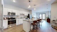 This thoughtfully designed one-bedroom, one-bathroom residence offers a cohesive open-concept layout with wood-style flooring flowing seamlessly throughout. The kitchen is a standout feature, showcasing crisp white shaker cabinetry, granite countertops, a full suite of stainless steel appliances, and a central island with pendant lighting overhead — perfect for both meal preparation and casual dining with barstool seating. The kitchen opens directly into the living area, which is bathed in natural light from a set of full-length French doors that provide outdoor views and enhance the airy ambiance of the space. The bedroom is a comfortable retreat with windows that invite natural light, complemented by a generously sized walk-in closet with wire shelving for ample storage. The bathroom features granite countertops consistent with the kitchen's design language, white cabinetry, and a well-lit vanity mirror. A dedicated utility room adds practical functionality to the layout. Neutral gray walls, clean lines, and consistent finishes throughout create a modern yet welcoming atmosphere that is both stylish and highly functional.