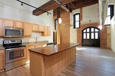 This single-floor loft-style space masterfully combines industrial heritage with contemporary living. The open main area makes a striking first impression with soaring ceilings showcasing exposed wooden beam rafters and visible ductwork, lending an authentic warehouse character. Rustic load-bearing wood columns punctuate the living space, while light hardwood flooring throughout adds warmth and continuity.

The kitchen is well-appointed with light oak cabinetry, granite countertops, and a full suite of stainless steel appliances including a refrigerator, electric range, built-in microwave, and dishwasher. A butcher-block breakfast bar provides additional workspace and casual dining. Track lighting mounted on the exposed beams ensures the kitchen and living areas are well lit.

A secondary room features original exposed brick walls running along the upper portion, paired with smooth drywall below and hardwood flooring, creating a dynamic mix of old and new. Natural light filters in through high windows, accentuating the textural contrast.

The bathroom is clean and functional, finished with large-format tile flooring, a light oak vanity with a single-basin sink and mirror, and a full soaking tub with shower combination. Dark wood doors throughout the unit provide a refined contrast to the lighter finishes, completing the space's cohesive industrial-modern aesthetic.