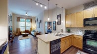 This single-floor residence offers a thoughtfully designed open-concept layout where the kitchen flows seamlessly into the living area. The kitchen is a functional centerpiece, featuring warm honey-toned cabinetry extending to the ceiling, granite-style countertops, a tile backsplash, and black appliances including a range, microwave, dishwasher, and refrigerator. A peninsula with bar seating accommodates multiple stools and is illuminated by a series of modern pendant lights, complemented by track lighting overhead. Dark hardwood-style flooring runs throughout the main living areas, adding warmth and continuity.

The living room benefits from large windows that invite natural light and feature draped curtains, while elevated ceilings enhance the sense of openness. A ceiling fan adds to the comfort of the space.

The bedroom is carpeted and well-lit by multiple windows with blinds and sheer curtains. It connects to a full bathroom finished with light tile flooring, a vanity with matching cabinetry, granite-style countertops, a soaking tub with a shower surround, and a large mirror. Adjacent to the bathroom is a generously sized walk-in closet with wire shelving, providing ample storage throughout.