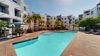 Located at 5395 Napa St in San Diego's Ocean Beach neighborhood, this outdoor amenity space showcases a stunning resort-style pool courtyard designed for relaxation and recreation under the Southern California sun. The centerpiece is a generously sized swimming pool with vivid turquoise water, complemented by a separate hot tub/spa that invites year-round enjoyment. The pool deck is finished in warm earth-toned concrete and features an ample lounge area equipped with chaise lounges, shaded seating beneath umbrellas, and covered pergola structures that provide comfortable respite from the sun. Lush palm trees and tropical plantings frame the space, enhancing the resort-like ambiance. The courtyard is enclosed by a multi-story building showcasing distinctive Spanish Colonial Revival architecture, characterized by crisp white stucco facades, arched accents, decorative rooflines, and private balconies that overlook the pool area. Glass-paneled fencing surrounds the pool, maintaining an open and airy feel while ensuring safety. The overall atmosphere is bright, welcoming, and ideally suited to the San Diego climate, offering residents a vibrant outdoor living experience in the heart of the city.