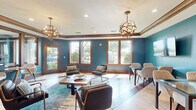This single-floor space presents a cohesive and well-appointed interior defined by bold teal walls contrasted against rich, warm-toned wood trim, baseboards, and door frames throughout. The main lounge area offers a comfortable arrangement of leather and upholstered accent chairs surrounding a rustic wood coffee table, all set atop a teal-toned area rug. Two striking globe pendant chandeliers with metallic finishes serve as focal points overhead, complemented by recessed lighting and decorative wood crown molding that adds architectural character to the ceiling line.

Large, wood-framed windows flood the space with natural light and offer views of the surrounding outdoor greenery and courtyard. A secondary dining area features glass-top tables paired with modern white and upholstered chairs, while wall-mounted flat-screen TVs make the space well-suited for presentations or group gatherings. Exposed wood ceiling beams further enhance the warm, layered aesthetic in this section.

The flooring throughout is a distinctive combination of light travertine tile and dark hardwood, laid in a geometric pattern that adds visual interest underfoot. Interior wood doors with paneled detailing maintain design consistency across the space, which also includes a corridor connecting to additional rooms. Overall, the space balances contemporary furnishings with classic architectural details, creating a welcoming and functional environment.