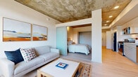 Located at 929 NE 26th Ave in Portland, Oregon, this one-bedroom, one-bathroom residence offers a thoughtfully designed layout that maximizes every square foot. A defining architectural feature throughout is the exposed concrete ceiling, which lends an industrial-modern character that contrasts beautifully with the warm light wood flooring found in every room.

The living area is bright and welcoming, anchored by floor-to-ceiling windows that invite abundant natural light and views of the surrounding neighborhood. The bedroom is separated from the living space by a frosted glass sliding door, providing privacy while maintaining an open, airy feel. The bedroom itself features the same exposed concrete ceiling and includes a dedicated closet with built-in shelving.

The galley-style kitchen is efficiently arranged with white cabinetry, stainless steel appliances including a gas range and dishwasher, a tile backsplash, and a clean white countertop — making excellent use of the available space. Built-in wall niches along the entryway corridor add a subtle decorative touch.

The bathroom is finished with modern gray cabinetry, a wide vanity with a white countertop, a large mirror, and direct access to a dedicated closet. Throughout the residence, a cohesive palette of white, light gray, and natural wood tones creates a calm, contemporary ambiance well-suited to urban living in Portland.