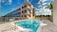 Located in Miami, FL, this outdoor pool deck offers a well-appointed communal amenity space designed for relaxation and recreation in a sunny South Florida setting. The centerpiece is a generously sized rectangular swimming pool with crystal-clear blue water, clearly marked depth indicators, and wide entry steps with stainless steel handrails for easy access. The pool is framed by a broad, tiled deck surface that provides ample space for lounging and movement around the perimeter.

The surrounding architecture is modern and clean, characterized by multi-story residential buildings with neutral warm-toned facades, large windows, and private balconies featuring sleek metal railings. The pool deck is enclosed by a metal security fence, ensuring a safe and defined outdoor environment. Utility access doors are set neatly into the building exterior, and a shower fixture is available poolside for convenience.

Lush palm trees add a tropical character to the space, complementing the vibrant Miami atmosphere. The open layout allows for abundant natural sunlight throughout the day, with wide open sky views creating a bright, airy ambiance. The combination of contemporary architectural design, thoughtful amenities, and Miami's iconic climate makes this pool deck an inviting outdoor retreat.