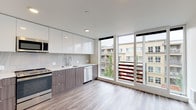 Located at 2100 East Madison Street in Seattle's Capitol Hill neighborhood, unit 503 is a contemporary single-level residence defined by clean lines and a modern aesthetic throughout. The open-concept layout seamlessly connects the kitchen and living areas, creating a bright, cohesive living environment.

The kitchen is a standout feature, showcasing high-gloss white upper cabinetry paired with wood-toned lower cabinets and sleek chrome hardware. Light quartz countertops and a tile backsplash complement the full suite of stainless steel appliances, including a range, built-in microwave, and dishwasher. Light wood-style flooring runs continuously throughout the unit, enhancing the sense of openness.

Floor-to-ceiling windows in both the kitchen and living areas flood the space with natural light and offer views of the surrounding neighborhood. The living area is generously proportioned, providing flexibility for various furniture arrangements.

The bathroom is finished in a minimalist style, featuring a wood-toned vanity with a white countertop, an undermount sink, a large mirror, a full soaking tub with shower, and wood-style flooring that ties back to the rest of the unit. The overall ambiance is clean, contemporary, and well-appointed.