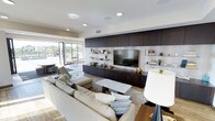 This single-floor space embodies contemporary design with a clean, open layout that feels both welcoming and sophisticated. The central living area is anchored by a generously sized sectional sofa in neutral tones, complemented by accent pillows and a stone-topped coffee table that adds a tactile, natural element to the refined interior.

The most striking architectural feature is the full-wall built-in media unit, crafted in rich dark wood cabinetry that extends from floor to ceiling. A large flat-screen television sits at its center, flanked by open glass shelving displaying tasteful decorative objects, books, and greenery. The contrast between the dark cabinetry and the light walls creates a bold, dramatic focal point that anchors the room.

Recessed ceiling lighting provides a soft, even illumination throughout, while a statement floor lamp adds a warm, ambient glow to the living space. Light hardwood flooring runs seamlessly through the area, contributing to the airy, open feel.

Large sliding glass doors and expansive windows along one wall flood the interior with natural light and offer views to the outdoors, creating a seamless connection between the interior and exterior. The overall ambiance is polished and modern, with a carefully curated palette of warm neutrals, dark wood tones, and natural materials throughout.
