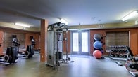 This single-level fitness center offers a functional and well-organized workout space designed to support a variety of exercise routines. The room features warm terracotta-toned accent walls paired with neutral ceiling tones, creating an energizing yet comfortable atmosphere. A cable resistance machine serves as a central strength-training fixture, accompanied by cardio equipment including an elliptical trainer visible along the side wall. A dedicated stability ball rack along the far wall holds multiple exercise balls of varying sizes, adding versatility for core and balance training. The space benefits from generous natural light streaming through a set of double glass doors, which open to what appears to be an outdoor balcony or terrace, enhancing ventilation and connectivity to the outdoors. The flooring is a smooth, easy-to-maintain surface appropriate for a fitness environment. Recessed lighting and flush-mounted fixtures provide consistent illumination throughout. The layout is open and spacious, allowing ample room for movement between equipment. An emergency exit sign above the glass doors reflects attention to safety standards. Overall, the space is thoughtfully arranged to accommodate both strength and functional fitness training.