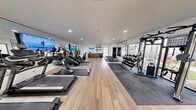 This fitness center offers a generously proportioned, single-floor exercise room designed to accommodate a comprehensive range of workout activities. The space features light-toned hardwood-style flooring throughout, complemented by crisp white walls and recessed lighting that create a bright, energizing atmosphere. Large floor-to-ceiling windows along multiple walls flood the interior with natural light and provide views of the surrounding outdoor environment, enhancing the open feel of the room.

The exercise room is outfitted with a robust selection of equipment, including multiple treadmills, elliptical machines, stair climbers, cable/multi-station strength machines, rowing machines, and a full dumbbell rack with a variety of free weights. Rubber floor mats are positioned beneath equipment stations for safety and durability. Large wall-mounted mirrors amplify the sense of space and allow users to monitor their form, while wall-mounted flat-screen televisions are integrated throughout the room for entertainment during workouts.

A built-in station along one wall includes shelving, a small refrigerator, and a water station for added convenience. A glass-paneled entry door provides easy access, and the overall layout is thoughtfully organized to allow efficient movement between cardio, strength, and stretching areas.