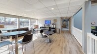 This single-floor fitness and wellness facility combines functional design with a welcoming atmosphere throughout its distinct zones. The social lounge area is bathed in natural light from expansive floor-to-ceiling windows that overlook outdoor courts, creating an airy and open feel. Light hardwood flooring runs throughout, complemented by white shiplap-style wall paneling that lends a clean, contemporary aesthetic. High-top tables and chairs are arranged throughout the lounge alongside a comfortable sofa seating area, making it an ideal spot for members to relax or socialize. A dedicated café station, branded with Starbucks, offers coffee and beverage service, complete with a wall-mounted display screen above. Magazine racks and wine displays add to the lifestyle-oriented atmosphere. A wide hallway connects the lounge to the rest of the facility, featuring recessed lighting, a drop ceiling, and a wall-mounted digital information display. The cardio fitness area is vibrant and energetic, featuring a bold orange accent wall lined with individual flat-screen monitors paired with a full row of ellipticals, stair climbers, and other cardio machines. White railing partitions define the space while maintaining an open sightline, and drop-panel ceiling lighting ensures the area is bright and well-lit throughout.