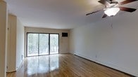 This single-floor space offers a clean, open layout characterized by smooth white walls and gleaming hardwood floors that reflect natural light throughout the interior. The main living area is bright and airy, anchored by a set of sliding glass doors that provide direct access to an outdoor balcony. The balcony overlooks a lush, tree-lined setting, bringing a sense of natural tranquility into the space and allowing for generous amounts of natural light to filter in. A ceiling fan is installed overhead, adding both functional air circulation and a subtle architectural detail to the room. Baseboard heating runs along the walls, ensuring efficient climate control throughout the space. The neutral white walls provide a versatile backdrop, making the space adaptable to a variety of interior design preferences. The overall ambiance is understated and practical, with an emphasis on clean lines and natural materials. The polished hardwood flooring is a standout element, adding warmth and a timeless quality to the interior. The connection to the outdoors through the sliding glass doors enhances the sense of openness and makes the space feel larger than its footprint suggests.