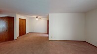 Located along US-12 E in Wayzata, Minnesota, this single-floor unit offers a practical and comfortable layout with consistent finishes throughout. The living area is generously proportioned, featuring wall-to-wall carpet, a textured ceiling, and a large sliding glass door dressed with vertical blinds that floods the room with natural light and provides outdoor views of the surrounding greenery. A wall-mounted heating unit keeps the space climate-controlled. An additional carpeted room, equipped with a ceiling fan and a closet with wood-toned doors, offers flexible use as a bedroom or secondary living space. The kitchen is outfitted with warm oak cabinetry, white countertops, a stainless steel sink, and includes a refrigerator, providing ample storage and a functional workspace. The bathroom features white subway tile wainscoting, a vanity with a marble-patterned countertop, an integrated sink, and a well-lit mirror framed by overhead globe lighting. Rich wood trim and warm-toned carpeting run throughout, creating a cohesive and inviting interior. The open layout, as seen from the overhead perspective, connects the main living space directly to the kitchen and hallway, offering an efficient and easy-to-navigate floor plan well suited to everyday living in the Wayzata area.