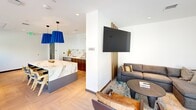 This single-floor space is thoughtfully designed with a contemporary aesthetic, combining functionality and refined style throughout its open layout. The interiors feature light herringbone-patterned hardwood flooring that runs consistently across the space, complementing the clean white walls and recessed lighting overhead.

A central highlight is the open lounge and social area, anchored by a large marble-topped island with rich walnut cabinetry below and a striking marble backsplash. Bold cobalt blue pendant lights hang overhead, adding a vibrant design accent. Adjacent to the kitchen area, a comfortable lounge zone features a sectional sofa, leather accent chairs, a wall-mounted flat-screen television, and warm wood slatted architectural details.

The dining area accommodates multiple high-top tables paired with natural wood cane-back chairs, lit by warm globe sconces along the walls. Horizontal band windows allow natural light to filter in while framing views of surrounding greenery.

Along the street-facing side, floor-to-ceiling glass windows and sliding panels flood an additional seating area with abundant natural light and urban street views. Wood panel wall accents with integrated sconces add warmth to this lounge nook. The overall ambiance is polished, welcoming, and well-appointed.