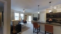 This well-appointed single-floor residence offers an open-concept layout that effortlessly connects the kitchen, dining, and living areas into one cohesive and inviting space. The living area is anchored by plush carpeting, a woven area rug, and comfortable seating arranged to create a relaxed and functional atmosphere. A ceiling fan with integrated lighting overhead adds both practicality and a finishing touch to the room. Large windows dressed with decorative curtains allow generous amounts of natural light to filter in, brightening the neutral-toned walls and creating an airy ambiance throughout. The kitchen features a raised breakfast bar with dark countertops and pendant lighting above, accompanied by upholstered bar stools that make it ideal for casual dining or entertaining. Dark cabinetry adds contrast and a modern edge to the kitchen's design. A private entry door at the rear of the living space provides additional access and light. Decorative wall art and tasteful accents are placed throughout, contributing to the warm and curated feel of the interior. The seamless flow between spaces and the abundance of natural light make this a comfortable and visually appealing home.