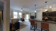 This well-appointed single-floor residence offers an open-concept layout that effortlessly connects the kitchen, dining, and living areas into one cohesive and inviting space. The living area is anchored by plush carpeting, a woven area rug, and comfortable seating arranged to create a relaxed and functional atmosphere. A ceiling fan with integrated lighting overhead adds both practicality and a finishing touch to the room. Large windows dressed with decorative curtains allow generous amounts of natural light to filter in, brightening the neutral-toned walls and creating an airy ambiance throughout. The kitchen features a raised breakfast bar with dark countertops and pendant lighting above, accompanied by upholstered bar stools that make it ideal for casual dining or entertaining. Dark cabinetry adds contrast and a modern edge to the kitchen's design. A private entry door at the rear of the living space provides additional access and light. Decorative wall art and tasteful accents are placed throughout, contributing to the warm and curated feel of the interior. The seamless flow between spaces and the abundance of natural light make this a comfortable and visually appealing home.