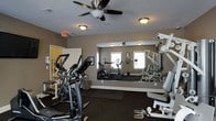 This single-floor fitness center is thoughtfully designed to provide a comprehensive workout environment. The space is outfitted with a variety of cardio equipment, including elliptical trainers and treadmills, arranged to allow comfortable movement between machines. A dedicated strength training area features a multi-station cable machine and a rack of free weights, catering to a range of exercise routines. Large wall-to-wall mirrors line one side of the room, visually expanding the space and allowing users to monitor their form during workouts. The dark rubberized flooring provides both durability and safety underfoot. Overhead, ceiling fans with integrated lighting fixtures ensure consistent air circulation and a well-lit environment throughout the workout area. Natural light filters in through multiple windows, contributing to an open and energizing atmosphere. The neutral taupe-toned walls give the space a clean, modern aesthetic. An emergency exit sign and clearly marked doorway ensure the space meets safety standards. Overall, this fitness center is a functional and well-appointed facility suited for a full range of exercise activities.