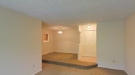 This single-floor space showcases a functional and straightforward layout with several notable architectural details. The living area features a sunken floor design, creating a subtle but distinct separation between the main living space and the entryway, adding visual interest to the open floor plan. Soft, wall-to-wall carpeting in a neutral tone extends throughout, contributing to a warm and comfortable atmosphere.

The textured popcorn ceiling adds a classic touch, while a pendant light fixture hangs centrally over the living area, providing focused ambient lighting. A small window along one wall allows for a modest amount of natural light to filter into the space.

The entryway is well-defined, featuring a ceiling-mounted flush light fixture and a set of bifold closet doors, offering convenient storage near the entrance. A decorative ledge or partial dividing wall separates the entry zone from the main living area, providing a sense of arrival without fully enclosing the space.

Overall, the layout is clean and open, with neutral tones throughout that create a blank canvas suitable for a variety of interior styles.