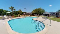 Located in Charleston, South Carolina, the Castlewood Townhomes community amenity space offers residents a welcoming outdoor retreat designed for relaxation and recreation. The centerpiece of this single-level outdoor area is a generously sized freeform swimming pool with crystal-clear blue water, complemented by a connected shallow wading section — ideal for all ages. The pool features multiple stainless steel entry rails for easy access, and the surrounding concrete deck provides ample space for lounge chairs and sunbathing. Mature palm trees and lush greenery frame the area, lending a tropical, resort-like atmosphere characteristic of the South Carolina Lowcountry. A covered gazebo is situated nearby, offering a shaded gathering spot for residents. Scattered lounge seating throughout the deck encourages leisurely outdoor living. The broader grounds are well-maintained, with open green space and additional seating visible in the background. Residential buildings of the community can be seen beyond the pool area, reinforcing the close-knit, neighborhood feel of the development. The space enjoys abundant natural sunlight, making it a vibrant and inviting hub for community life throughout the warmer months.