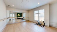Located at 601 W Bacon St in Richmond, VA, this single-room fitness and barre studio is thoughtfully designed to maximize functionality and ambiance within a compact footprint. The space is anchored by a wall of floor-to-ceiling mirrors that visually expands the room, paired with a wall-mounted wooden ballet barre that spans the length of the mirrored wall — ideal for barre workouts, stretching routines, and dance practice. Smooth, light-toned hardwood flooring runs throughout, providing a clean, resilient surface well-suited for a variety of fitness activities. Natural light flows in through a large multi-pane window, lending the studio a bright, open feel. A standout decorative feature is a lush, full-coverage living green wall adorned with a glowing neon sign reading "THIS MUST BE THE PLACE," adding a bold, welcoming personality to the entry area. A warm-toned curved bench provides a functional seating accent near the entrance. Recessed ceiling lighting supplements the natural light, ensuring the studio remains well-illuminated. Exercise equipment, including a fitness machine, is also present within the space. The overall design blends a clean, modern aesthetic with motivating, character-rich details, creating an inviting environment for fitness and wellness activities.