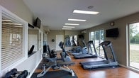 This fitness center offers a comprehensive workout environment designed to accommodate a range of exercise needs. The space features warm hardwood flooring accented by rubber equipment mats, providing both an inviting aesthetic and a practical, durable surface for training. Overhead, a series of recessed rectangular panel lights ensure bright, even illumination throughout the room, complementing the natural light that streams in through large sliding glass windows along one wall, offering pleasant views of the surrounding greenery outdoors. The cardio equipment lineup includes multiple treadmills, elliptical machines, and a stationary bike, with brands such as Vision Fitness and Hoist represented. A dedicated strength training bench adds versatility for resistance workouts. Wall-mounted televisions are positioned for easy viewing during exercise sessions. A mirrored wall section toward the rear of the space enhances the sense of depth while allowing users to monitor their form. A wall-mounted mini-split air conditioning unit ensures comfortable temperature control year-round. The neutral-toned walls and clean, organized layout contribute to a focused and functional atmosphere, making this fitness center a practical and well-appointed facility for daily exercise routines.