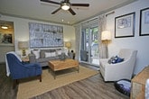 This single-floor residence offers a comfortable and thoughtfully designed living space with a warm, inviting ambiance. The living room serves as the centerpiece of the home, characterized by smooth ceilings adorned with a brushed nickel ceiling fan with integrated lighting, and crown molding that adds a refined architectural touch. Rich hardwood-style flooring anchors the space, complementing the neutral wall tones throughout.

Natural light fills the room through a large sliding glass door dressed with elegant patterned drapes, which opens directly onto a private balcony — an ideal spot for outdoor relaxation with views of surrounding greenery. The living area is styled with a plush gray sofa, a classic accent chair in crisp white upholstery, and a warm wood media console, all centered around a woven area rug and a geometric coffee table.

Wall art in black frames adds a contemporary decorative element, while coordinating floor and table lamps provide layered lighting options. The overall design balances modern sensibility with everyday comfort, making the space feel both functional and aesthetically cohesive. The single-floor layout ensures seamless flow and accessibility throughout the residence.