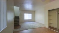 This two-story residence offers a straightforward and functional layout designed to maximize usable living space across both levels. The main floor features an open living area that combines carpet and hardwood-style flooring, creating a clear distinction between relaxation and transitional zones. A large window fitted with vertical blinds allows natural light to fill the room, contributing to a bright and airy atmosphere. The textured ceiling adds a subtle architectural detail to the otherwise clean, neutral-toned interior. A carpeted staircase positioned along one wall provides access to the second floor, integrating seamlessly into the living space without disrupting the open feel of the room. The walls are finished in a soft, neutral palette that offers a versatile backdrop suitable for a variety of interior styles. The overall design is practical and uncluttered, emphasizing open space and ease of movement. The two-floor configuration provides natural separation between common living areas and the upper level, offering a sense of privacy and organization throughout the home.