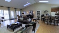 This thoughtfully designed single-floor space makes an immediate impression with its dramatic vaulted ceilings and multiple skylights that flood the interior with an abundance of natural light. The main gathering area is generously proportioned, centered around a comfortable seating arrangement featuring plush, dark upholstered sofas and armchairs positioned atop a patterned area rug over gleaming hardwood floors. A stylish coffee table and side tables anchor the lounge area, creating an inviting setting for relaxation or conversation. Towards one end of the space, a sophisticated bar-height seating area provides an additional social zone, complete with counter stools and accent lighting. A decorative mirrored cabinet serves as a striking focal point along the back wall, flanked by tasteful table lamps and elegant floral arrangements. Large windows dressed with soft draping curtains frame views of the exterior, enhancing the sense of openness and connection to the outdoors. The warm, neutral color palette of soft beige walls paired with rich wood tones and deep charcoal furnishings creates a cohesive and refined ambiance. This space is well-suited for gatherings, socializing, or simply unwinding in a polished and comfortable environment.