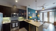 This well-appointed single-story unit in Houston, Texas, offers a thoughtfully designed open-concept layout that seamlessly connects the kitchen, dining, and living areas. The kitchen is a standout feature, showcasing rich dark cabinetry with glass-front uppers, a subway tile backsplash, stainless steel appliances including a range, microwave, and dishwasher, and a peninsula with a double sink and bar seating — ideal for casual dining or entertaining. Pendant lighting and track lighting illuminate the space effectively. The living area is anchored by a bold teal accent wall, large windows with blinds and curtains that invite natural light, and a ceiling fan overhead. A built-in desk nook with dark upper cabinetry provides a functional workspace tucked conveniently off the main living area. The bedroom is generously sized, featuring plush carpeting, large windows, a ceiling fan, and ample room for full-sized furniture. The bathroom offers a dual-sink vanity with a wide frameless mirror, dark cabinetry, light countertops, and a glass-enclosed shower with white subway tile. Dark hardwood-style flooring runs throughout the main living areas, adding warmth and continuity to the overall design.