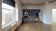 This single-floor space showcases a loft-inspired design that blends industrial character with modern finishes. The open main living area is anchored by polished concrete floors that reflect natural light, contributing to a bright and airy atmosphere. Exposed concrete beams traverse the ceiling, adding architectural interest and reinforcing the space's industrial aesthetic. Large windows along the perimeter frame urban skyline views and flood the interior with generous natural light, while classic cast-iron radiators underscore the building's historic character.

The kitchen is seamlessly integrated into the open living area and serves as a visual focal point, featuring a striking deep-blue subway tile backsplash that extends from counter to ceiling. Stainless steel appliances — including a gas range with an overhead ventilation hood, dishwasher, and refrigerator — are complemented by gray cabinetry and open brass-toned wall shelving, creating a contemporary yet functional cooking space.

The bathroom continues the modern design language with a wall-mounted navy vanity, a mirrored medicine cabinet, industrial-style sconce lighting, and a full tub-shower combination, all finished in clean white tones.