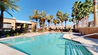 This inviting outdoor amenity space showcases a resort-style setting defined by its expansive swimming pool with crystal-clear turquoise water, complemented by a bubbling hot tub spa for relaxation. The pool area is generously sized, offering ample room for recreation and leisure, with wide concrete pool decking that provides comfortable space for lounging and movement around the water's edge.

Towering fan palms and date palms frame the space dramatically, creating a lush, tropical ambiance that contrasts beautifully with the warm desert surroundings. The landscaping throughout the area features a thoughtful mix of desert-adapted plants, shrubs, and ground-level greenery, adding color and texture to the hardscaped surfaces.

The surrounding buildings exhibit a Southwestern architectural style, characterized by warm earth-tone stucco exteriors in shades of terracotta and tan, with clean geometric lines and multi-level balconies accented by wrought iron railings. A dedicated clubhouse or recreational facility is visible adjacent to the pool, offering additional amenity space for residents.

The overall ambiance is one of a sun-soaked desert oasis — bright, open, and welcoming — with the brilliant blue sky serving as a natural backdrop. This outdoor space is clearly designed to maximize the enjoyment of a warm-weather climate, offering both active and relaxing options in a beautifully maintained setting.