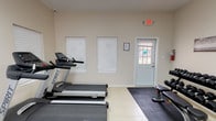 This fitness center is a fully equipped, single-floor facility designed to support a comprehensive workout routine. The space features light-toned walls, large format tile flooring, and protective rubber matting strategically placed beneath equipment zones, creating clearly defined areas for different types of exercise.

The cardio section is anchored by two Spirit treadmills positioned side by side, complemented by an elliptical trainer and a recumbent stationary bike, offering a well-rounded range of low- and high-impact options. A multi-function cable resistance machine provides a versatile strength training station capable of supporting a wide variety of exercises.

The free weights area includes a fully stocked three-tier dumbbell rack with a complete set of rubber hex dumbbells and an adjustable workout bench, catering to users of all fitness levels. A water cooler station is conveniently located within the space, ensuring hydration is easily accessible during workouts.

Natural light filters in through multiple windows fitted with privacy blinds, contributing to a bright and open atmosphere. Overhead fluorescent lighting ensures consistent illumination throughout. Safety is addressed with clearly marked exit signage and a fire extinguisher mounted on the wall. Decorative wall art adds a finishing touch to the functional environment.