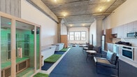 This single-floor space in Alameda, California, blends industrial architecture with modern functionality across a generous footprint. Exposed concrete ceilings with structural columns and beams define the aesthetic throughout, complemented by polished concrete floors in the main living areas and large multi-pane windows that flood the interior with natural light and offer open views of the surrounding neighborhood.

The open-plan kitchen and living area features warm wood cabinetry, subway tile backsplash, stainless steel appliances including a full-size refrigerator, oven, microwave, and dishwasher, alongside in-unit washer and dryer. The living area accommodates comfortable seating arrangements beneath high ceilings, lending an airy, loft-like atmosphere.

A dedicated wellness zone is a standout feature, equipped with an infrared sauna, cold plunge tubs set on turf runners, and ample open floor space suitable for yoga or fitness use, all beneath the signature exposed concrete ceiling. A lounge area with upholstered seating completes this section.

Two bathrooms are finished in a clean, contemporary style with subway tile shower surrounds, patterned tile flooring, and modern fixtures, providing functional and polished amenities throughout the space.