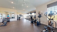 Located at 6160 Hall Rd in Galloway, Ohio, this fitness center offers a comprehensive and thoughtfully arranged exercise facility within a generously sized single-floor space. The room is bathed in natural light courtesy of multiple windows dressed with patterned Roman shades, providing views of the surrounding outdoor environment while maintaining a bright and energizing atmosphere. Recessed lighting and a ceiling fan supplement the natural light, ensuring the space remains well-lit and comfortable during workouts.

The cardio section is prominently featured, housing a row of treadmills, upright and recumbent stationary bikes, and elliptical cross-trainers — all positioned to take advantage of the natural light and mounted flat-screen televisions. The strength and functional training area is equally well-appointed, offering a cable machine, a rowing machine, a weight bench, a dumbbell rack, and an assortment of stability and medicine balls for dynamic exercises.

Full-length mirrors line portions of the walls, visually expanding the space and allowing users to monitor their form throughout their workouts. The durable rubber flooring provides both safety and comfort underfoot. With its clean, neutral color palette of warm beige walls and white trim, the fitness center maintains an inviting and professional ambiance suitable for all fitness levels.