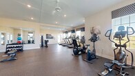 Located at 6160 Hall Rd in Galloway, Ohio, this fitness center offers a comprehensive and thoughtfully arranged exercise facility within a generously sized single-floor space. The room is bathed in natural light courtesy of multiple windows dressed with patterned Roman shades, providing views of the surrounding outdoor environment while maintaining a bright and energizing atmosphere. Recessed lighting and a ceiling fan supplement the natural light, ensuring the space remains well-lit and comfortable during workouts.

The cardio section is prominently featured, housing a row of treadmills, upright and recumbent stationary bikes, and elliptical cross-trainers — all positioned to take advantage of the natural light and mounted flat-screen televisions. The strength and functional training area is equally well-appointed, offering a cable machine, a rowing machine, a weight bench, a dumbbell rack, and an assortment of stability and medicine balls for dynamic exercises.

Full-length mirrors line portions of the walls, visually expanding the space and allowing users to monitor their form throughout their workouts. The durable rubber flooring provides both safety and comfort underfoot. With its clean, neutral color palette of warm beige walls and white trim, the fitness center maintains an inviting and professional ambiance suitable for all fitness levels.