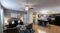 This single-floor residence showcases an open-concept layout that seamlessly connects the living area to the kitchen, creating a spacious and cohesive environment. The living room features warm hardwood-style flooring, neutral gray accent walls, and a ceiling fan with integrated lighting, contributing to a comfortable and well-lit atmosphere. A large piece of wall art adds a urban, contemporary touch to the space.

The kitchen is outfitted with dark cabinetry, a contrasting light countertop, and a peninsula with bar-height seating, making it ideal for casual dining or entertaining. Pendant lighting hangs above the peninsula, adding both style and functional illumination. Dark-finish appliances complement the overall modern aesthetic.

Visible through a hallway, an in-unit laundry area provides added convenience. The open corridor design connects the main living spaces efficiently while maintaining a sense of openness. Throughout the unit, consistent hardwood-style flooring unifies the space. The overall ambiance is modern and urban, with a carefully coordinated color palette of grays, blacks, and teal accents.