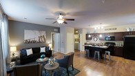 This single-floor residence showcases an open-concept layout that seamlessly connects the living area to the kitchen, creating a spacious and cohesive environment. The living room features warm hardwood-style flooring, neutral gray accent walls, and a ceiling fan with integrated lighting, contributing to a comfortable and well-lit atmosphere. A large piece of wall art adds a urban, contemporary touch to the space.

The kitchen is outfitted with dark cabinetry, a contrasting light countertop, and a peninsula with bar-height seating, making it ideal for casual dining or entertaining. Pendant lighting hangs above the peninsula, adding both style and functional illumination. Dark-finish appliances complement the overall modern aesthetic.

Visible through a hallway, an in-unit laundry area provides added convenience. The open corridor design connects the main living spaces efficiently while maintaining a sense of openness. Throughout the unit, consistent hardwood-style flooring unifies the space. The overall ambiance is modern and urban, with a carefully coordinated color palette of grays, blacks, and teal accents.