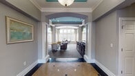 Located at 2400 Garden Hill Drive in Raleigh, NC, this single-story community clubhouse presents a refined and welcoming environment defined by thoughtful architectural details and upscale finishes throughout. The grand entrance foyer sets the tone immediately, featuring polished terrazzo flooring with decorative inlay, tray ceilings with teal accents, bold crown molding, and arched transom windows flanking the front door that flood the space with natural light. A large-scale artwork anchors the entry wall, contributing to an atmosphere of sophistication. Beyond the foyer, a wide arched passageway flanked by decorative columns leads into a spacious lounge and gathering area. This central space combines a seating area with plush furnishings and a bar-height counter, all positioned beneath a striking teal barrel-vaulted ceiling detail. Adjacent to the main lounge, a sunlit nook features soaring floor-to-ceiling windows with a curved bay configuration, offering views of the outdoor pool area and filling the room with abundant natural light. Stylish patterned carpet, layered drapery in coordinating teal and gold tones, and contemporary furnishings enhance the area's inviting character. A compact, well-appointed kitchenette includes dark cabinetry, granite countertops, stainless steel appliances, and wood-look flooring. Additional seating alcoves with teal upholstered chairs and glass-top tables provide comfortable, versatile gathering spots throughout the space.